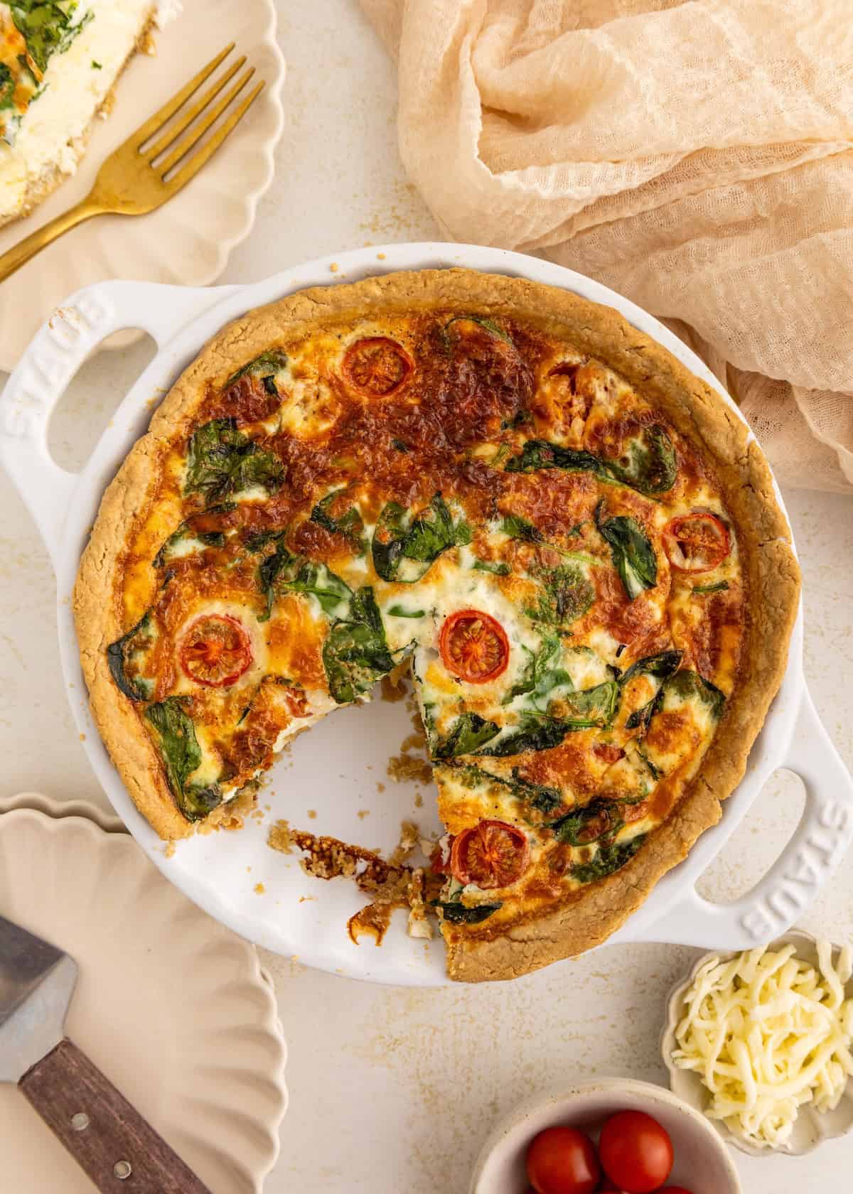 Quiche with spinach and tomatoes in a white dish, missing one slice, on a light table.