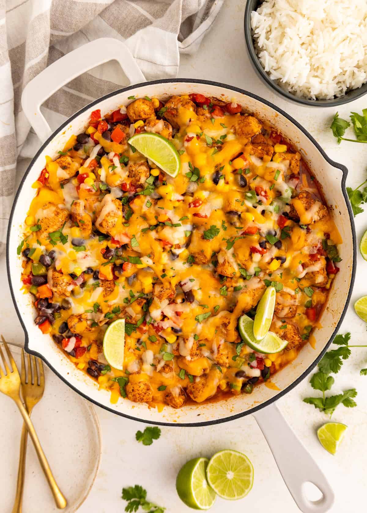 A skillet of cheesy chicken and veggies with lime wedges, served next to a bowl of rice.