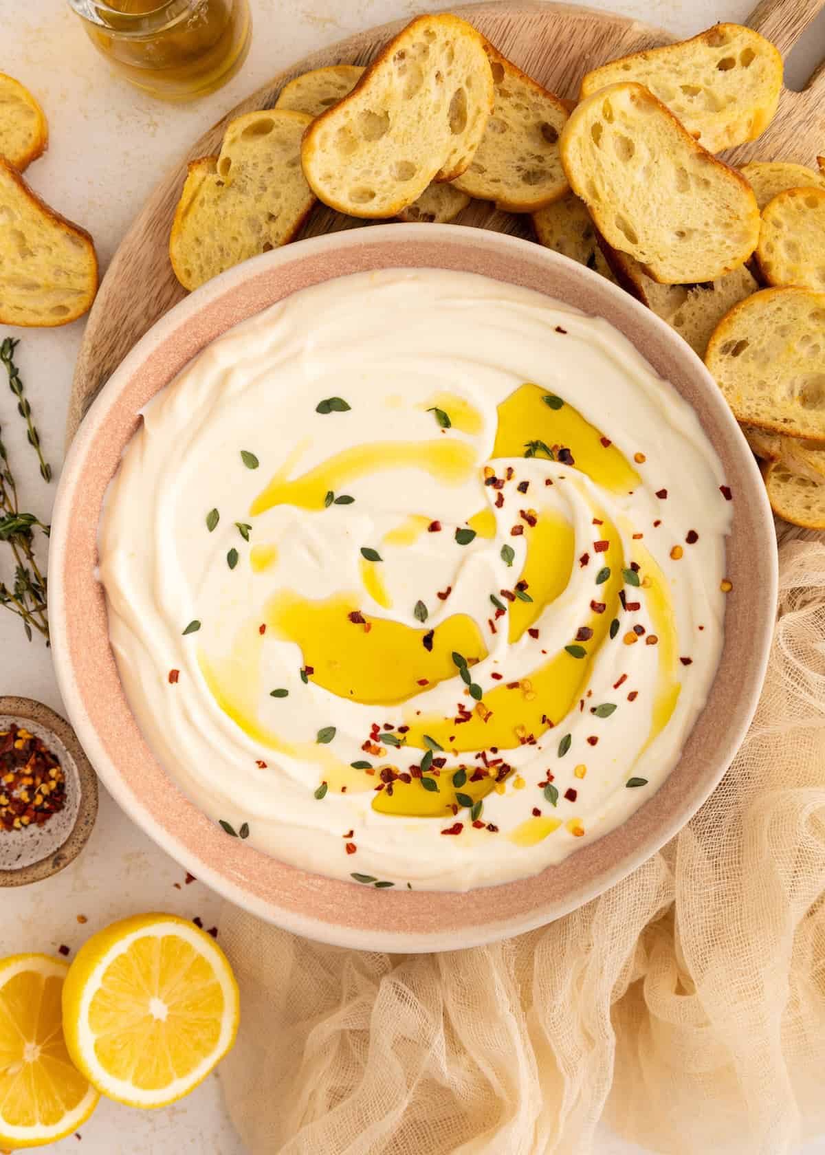 Creamy dip topped with olive oil, herbs, and chili flakes, surrounded by toasted bread slices.