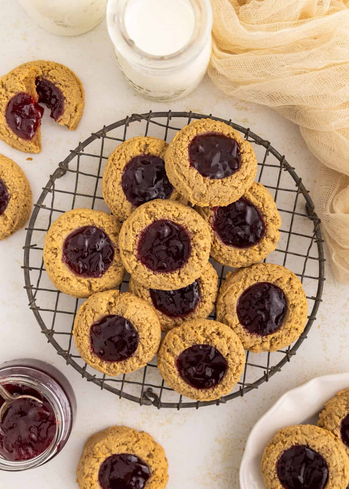 Gluten-free thumbprint cookies with jam cooling on a rack, milk and jam jars nearby.