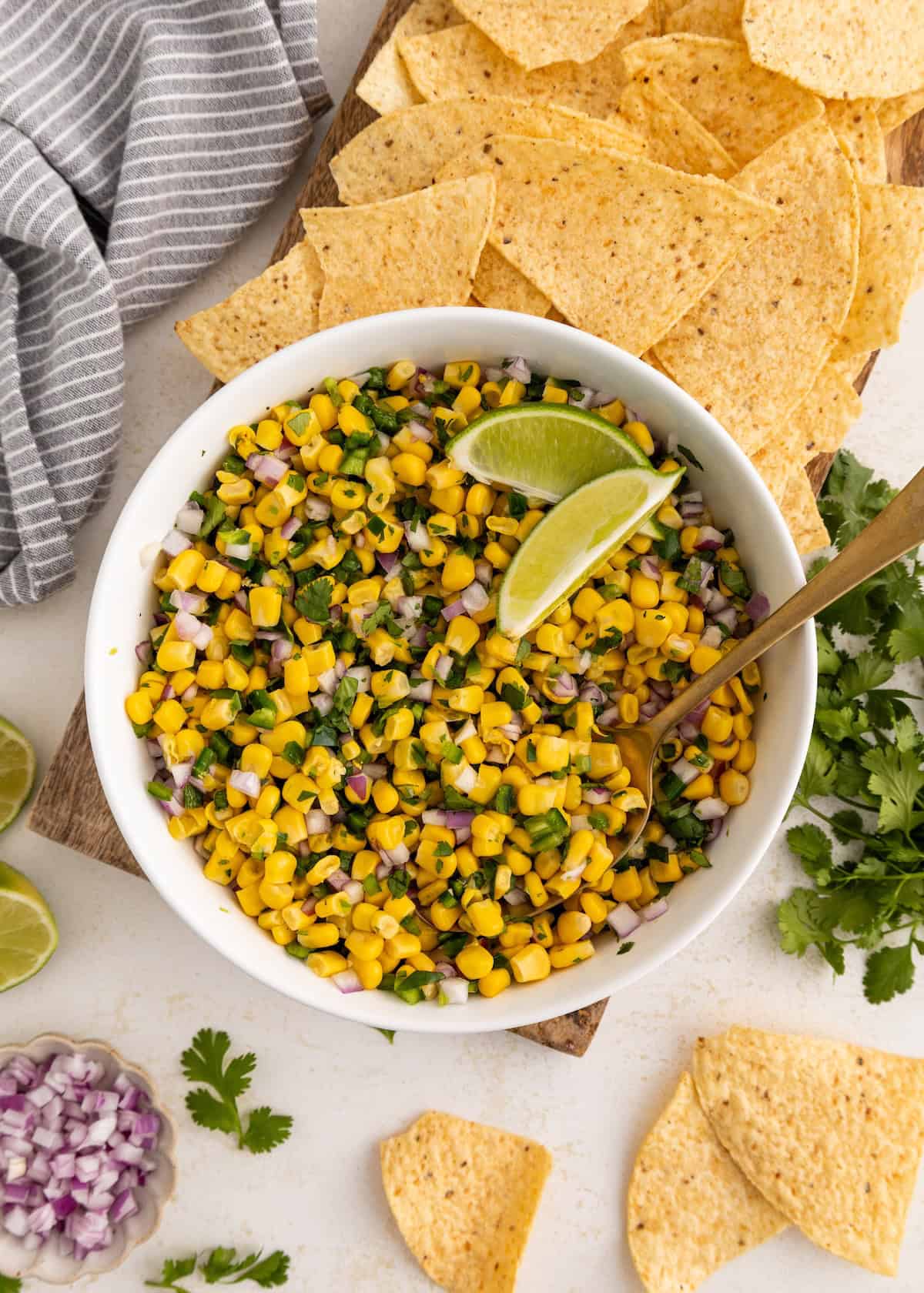 Bowl of corn salsa with lime wedges, surrounded by tortilla chips and fresh cilantro.